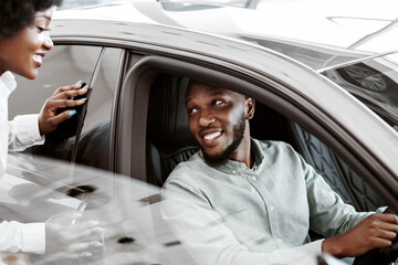 Test drive. Cheerful black man sitting in driver seat of new car, choosing vehicle with his wife...