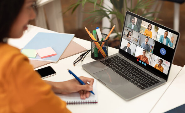 A woman sits at a desk with a laptop open, displaying a video conference meeting with several participants. She is taking notes in a notebook with a blue pen, her focus on the screen.