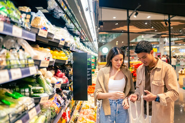 Happy Asian family couple using reusable cotton shopping bag buying food at supermarket. Man and woman enjoy sustainable living and healthy lifestyle choosing organic vegetable and fruit on shelves.