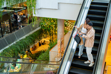 Happy Asian family man and woman shopping and buying goods at department store. Couple enjoy urban lifestyle holding shopping bag standing on escalator in shopping mall on summer holiday vacation.