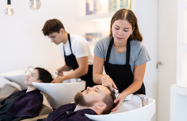 Fototapeta premium Young woman stylist washes male clients hair, prepares it for cutting and styling. Barbershop visitor placed head in sink, enjoying process of hair care