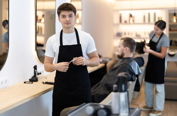 Obraz premium Young man hairdressers stylist rests during break, in absence of clients. Advertising poster for chain of hairdressing salons. Employee is waiting for arrival of client with scissors in hands.