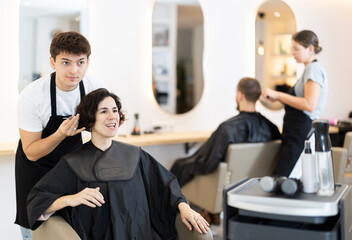 Young guy hairdresser stylist gives haircut to adult woman client in beauty salon