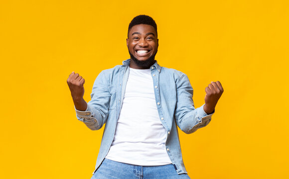 Portrait of overjoyed black man celebrating success with clenched fists and euphoric face expression on yellow studio background with copy space