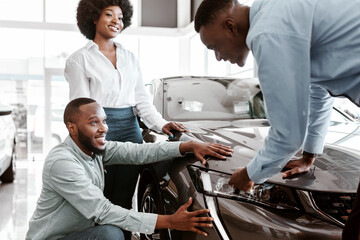 Salesman talking to young black couple, showing them new luxury car at auto dealership. African american clients buying or renting automobile at showroom. Vehicle local distribution concept