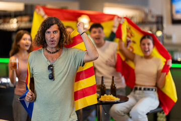 Group of friends fans watching match cheering with flags of Catalonia and Spain in bar