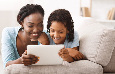 Fototapeta premium African Mom And Daughter Watching Cartoons Together On Digital Tablet, Lying On Couch. Free space