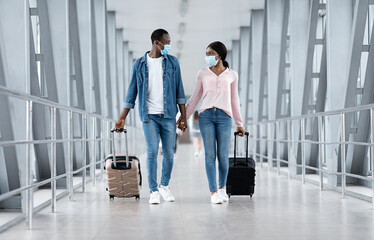 Coronovirus Travels. Black couple in masks walking with luggage at airport building, holding hands and looking at each other, copy space