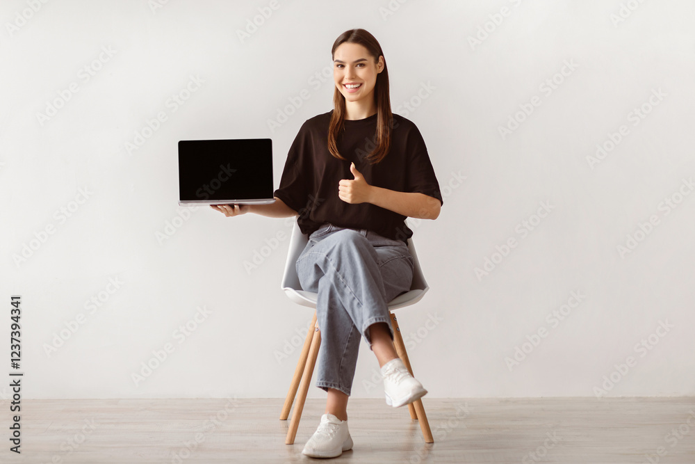 Poster smiling young woman sitting on chair, showing laptop with empty screen, gesturing thumb up, mockup f