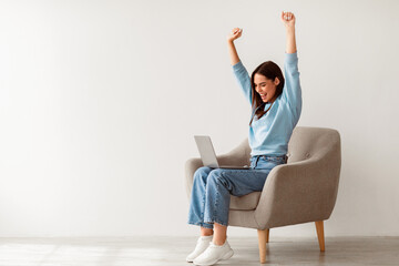 Overjoyed millennial woman sitting in armchair with laptop, celebrating huge online win or success against white studio wall, copy space. Young lady enjoying big sale in web store