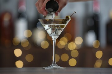 Woman making tasty martini cocktail with olives at wooden table on blurred background, closeup. Bokeh effect