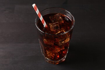 Refreshing cola with ice cubes and drinking straw in glass on black wooden table, closeup
