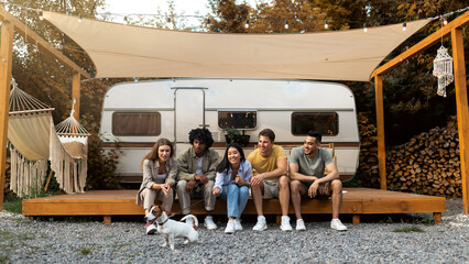 Group of cheery young diverse people with their cute dog sitting near motorhome, having fun camping trip in autumn. Multiethnic millennial friends relaxing together on weekend in countryside © Prostock-studio