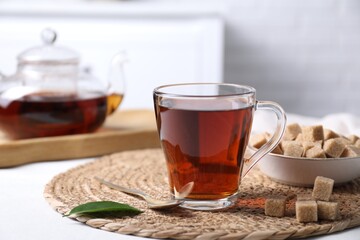 Aromatic black tea in cup and brown sugar cubes on white table, closeup