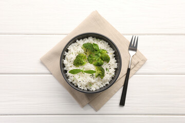 Tasty rice with broccoli and basil in bowl on white wooden table, top view
