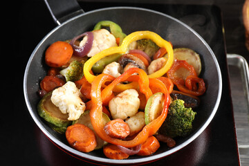 Frying pan with vegetables and stove on table against black background, closeup
