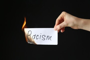 Woman holding burning card with word Racism on black background, closeup