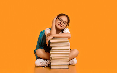Cute Korean Schoolgirl Sitting At Books Stack In Studio Over Yellow Background, Looking At Camera. School Education And Homework