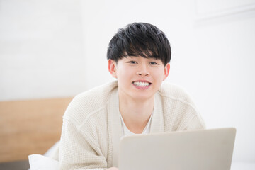 Young smiling man in his 20s lying face down on the bed and looking at the camera while he is on the computer. Image of a young student or freelance worker.