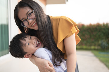 Happy Thai mother hugging her laughing son outside the house, sharing a joyful and loving moment...