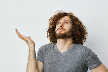 Thoughtful young man with curly hair contemplating in a gray shirt against a neutral background,...
