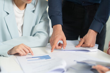 A woman in a light blazer reviews documents while another person points at the paperwork,...