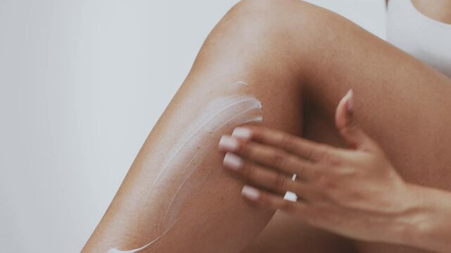 A millennial African American woman is enjoying a self-care moment at home, applying a skin moisturizer on her leg to nourish and pamper her body. The atmosphere is serene and focused.