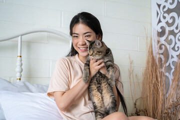 Obraz premium Smiling young Asian woman holding her adorable tabby cat in a cozy bedroom environment, looking at camera