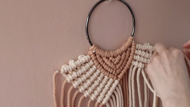 Hands carefully crafting a macrame wall hanging with beige and brown cords. Bohemian-inspired decor coming to life, highlighting the art of handmade design and the creative DIY process.