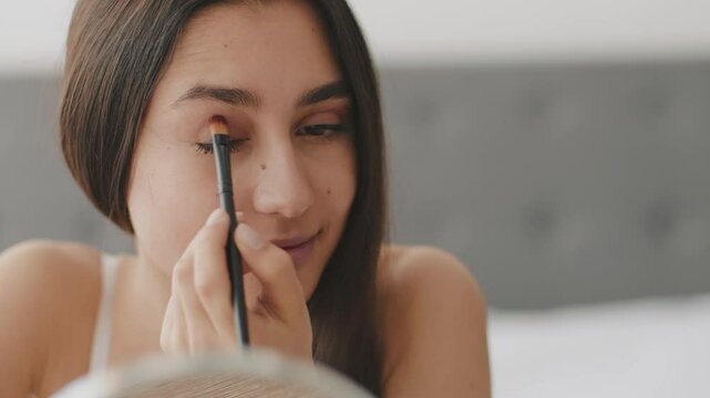 A young Middle Eastern woman engages in her skincare and makeup regimen at home. She delicately applies eyeshadow while looking into a mirror, highlighting her dedication to self-care.