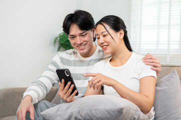 A happy young Asian couple sits together on a sofa in the living room, watching a video on a phone.