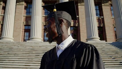 An African American student proudly smiles while wearing a graduation cap and black gown on the steps of a grand university building. High education concept