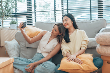 Fototapeta premium Two young Asian women smiling and happy while taking a selfie in their living room at home, looking at the camera
