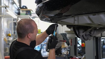An auto mechanic working under the car on a car lift in the garage, he disassembles the bottom of the car for further repair. Antifreeze drain in the service station. Auto repair shop, auto repair.