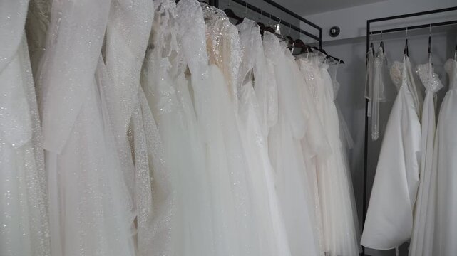 Many different wedding dresses are hanging on hangers in a modern wedding salon. Various wedding dresses. Wedding fashion. Stable movement of the camera backwards. Slow motion.