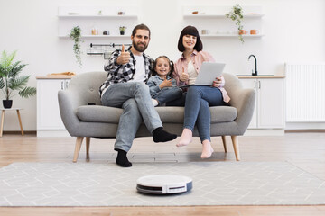 Caucasian family with parents and daughter relaxing on sofa in modern living room with robotic vacuum. Father, mother, and child showing thumbs up. Home technology and bonding scene.