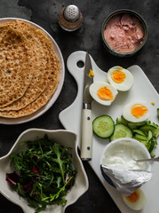 Ingredients for cooking  crepes stuffed with tuna, cucumber, eggs, arugula salad on a dark background, top view