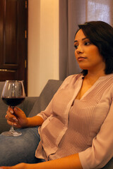 Woman relaxing at home with glass of red wine, wearing casual blouse while sitting on comfortable couch in warm evening atmosphere