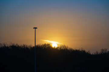Fototapeta premium Dramatic winter sunrise behind silhouetted treeline with tall floodlight pole in the foreground. Golden morning sun breaking through the dark sky creates an atmospheric British landscape