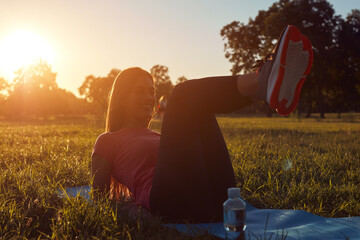 Fototapeta premium Young woman exercising on a sports yoga mat on the grass in urban park during summertime sunset time.
