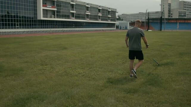 Medium rear shot of Caucasian male para athlete with acheiria disability training for competition at stadium, throwing javelin far then walking over green grass lawn to retrieve it