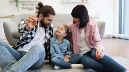 Caucasian family indoors excited after moving to new home. Parents and child smiling while holding house keys. Young couple with daughter celebrating. Casual clothing.
