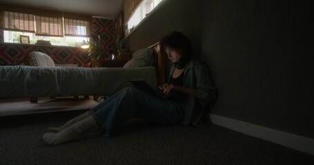 A young woman uses a laptop sitting on the floor in the bedroom at home with a serious look