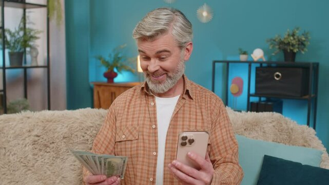 Mature man at home holding smartphone and money dollar cash while making financial transfer through mobile app. Middle-aged guy happy from quick transaction, pleased with his earnings, salary, savings