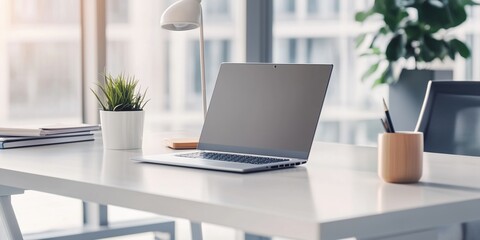 Fototapeta premium Modern white desk setup with laptop, coffee cup, and potted plant in a bright office space. concept of productivity and minimalist workspace