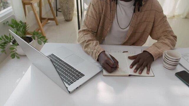 Cropped shot of unrecognizable African American office employee doing project task on wireless laptop and taking notes when sitting at desk in cozy office space