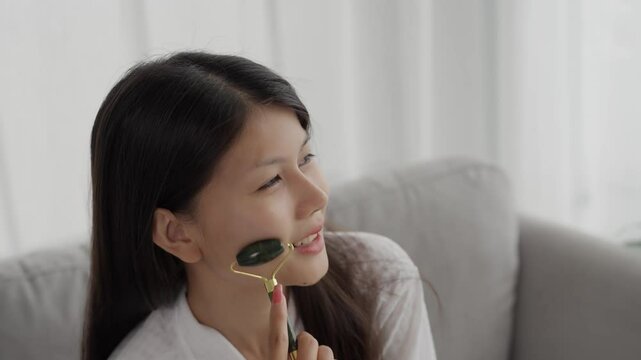 Asian woman in bathrobeses gua sha quarz lifting tool on face. Woman self massage with jade gouache scraper
