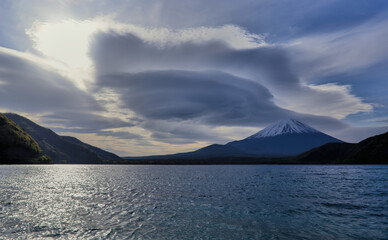 Morning view of a snow-capped Mount Fuji from the north shore of Lake Motosu, with a unique circular cloud formation partially blocking the sun