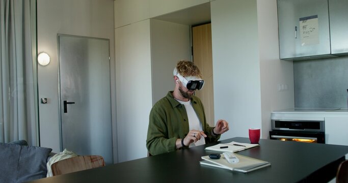 A man in a virtual reality headset typing on a virtual keyboard sitting at a table at home. Daylight