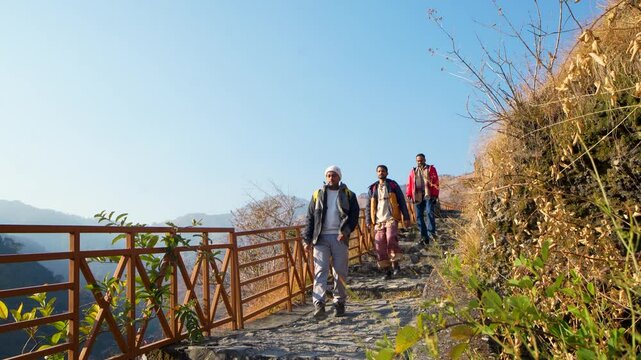 Three men hiking on a mountain trail in a row, all smiling and enjoying the journey. The background features the scenic Tiger Fall and the Kanaser Range in Chakrata, Uttarakhand, India.